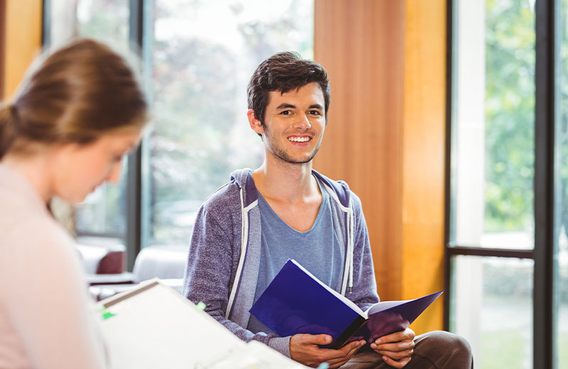 A student with a smiling face is holding a blue notebook